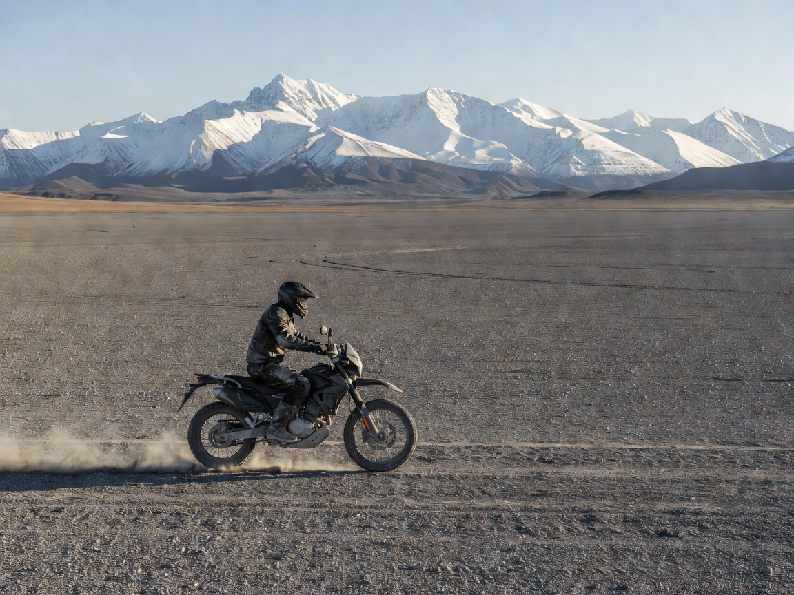 Motorcyclist riding on a vast gravel plain with snow-capped mountains in the background